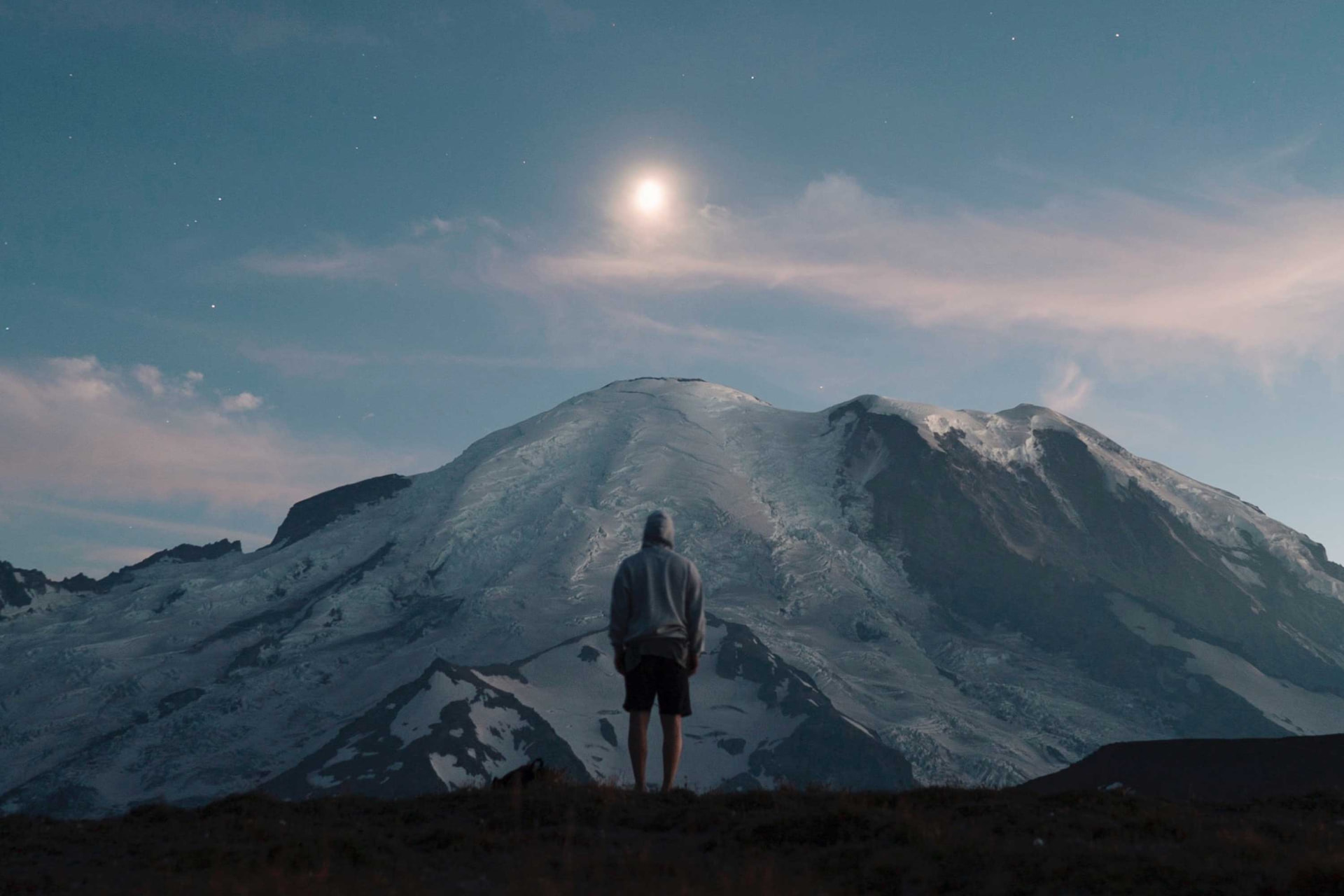 A person standing before a moonlit mountain range at dusk