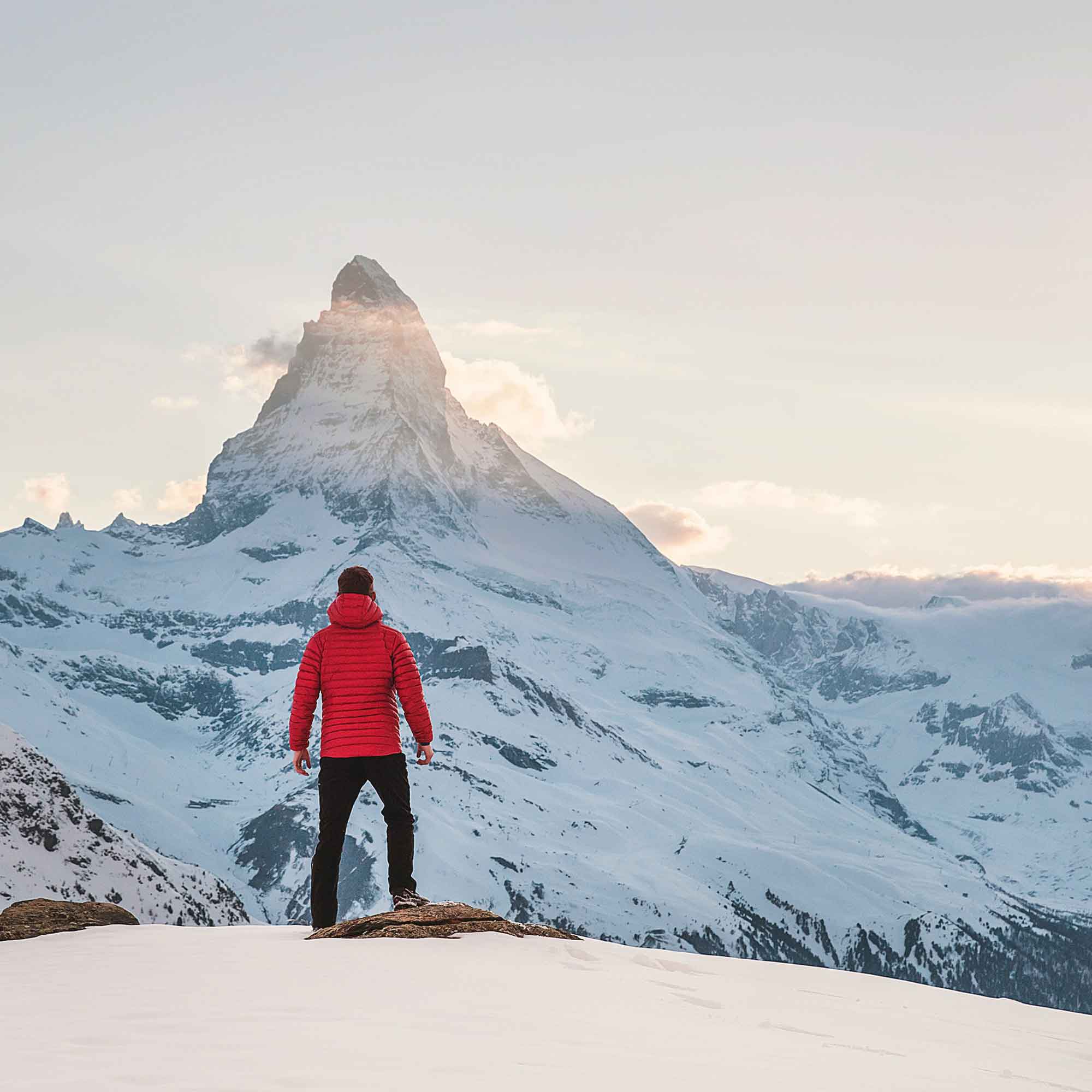 A person in a red jacket looking towards a snow-covered mountain peak